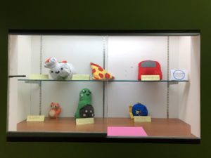 Display case with sewn objects, including a stuffed sushi, avocado, and pizza