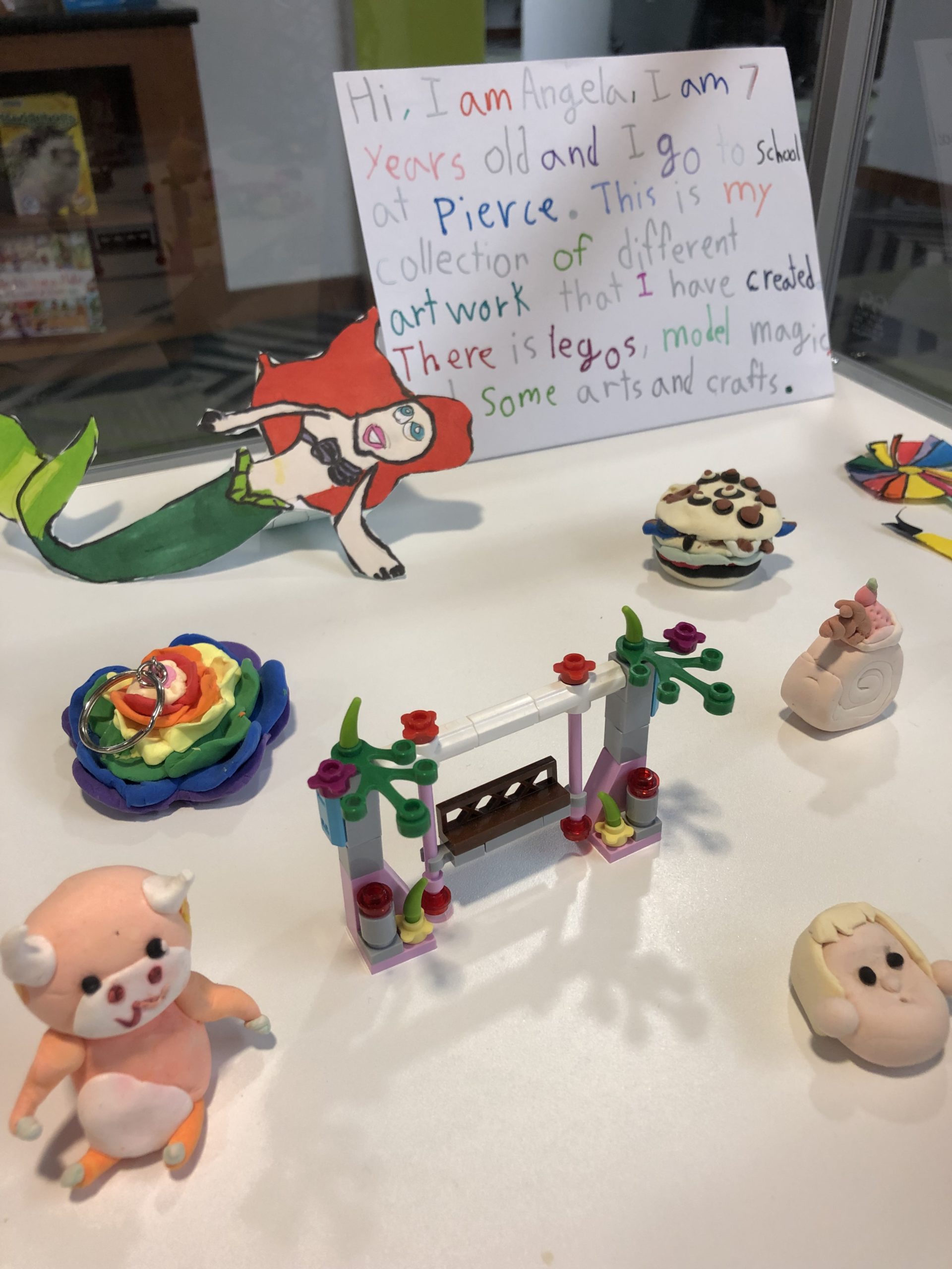 Display of small craft projects with a hand-written sign that reads "Hi, I am Angela, I am 7 years old and I go to school at Pierce. This is my collection of different artwork that I have created. There is Legos, Model Magic, and some arts and crafts."
