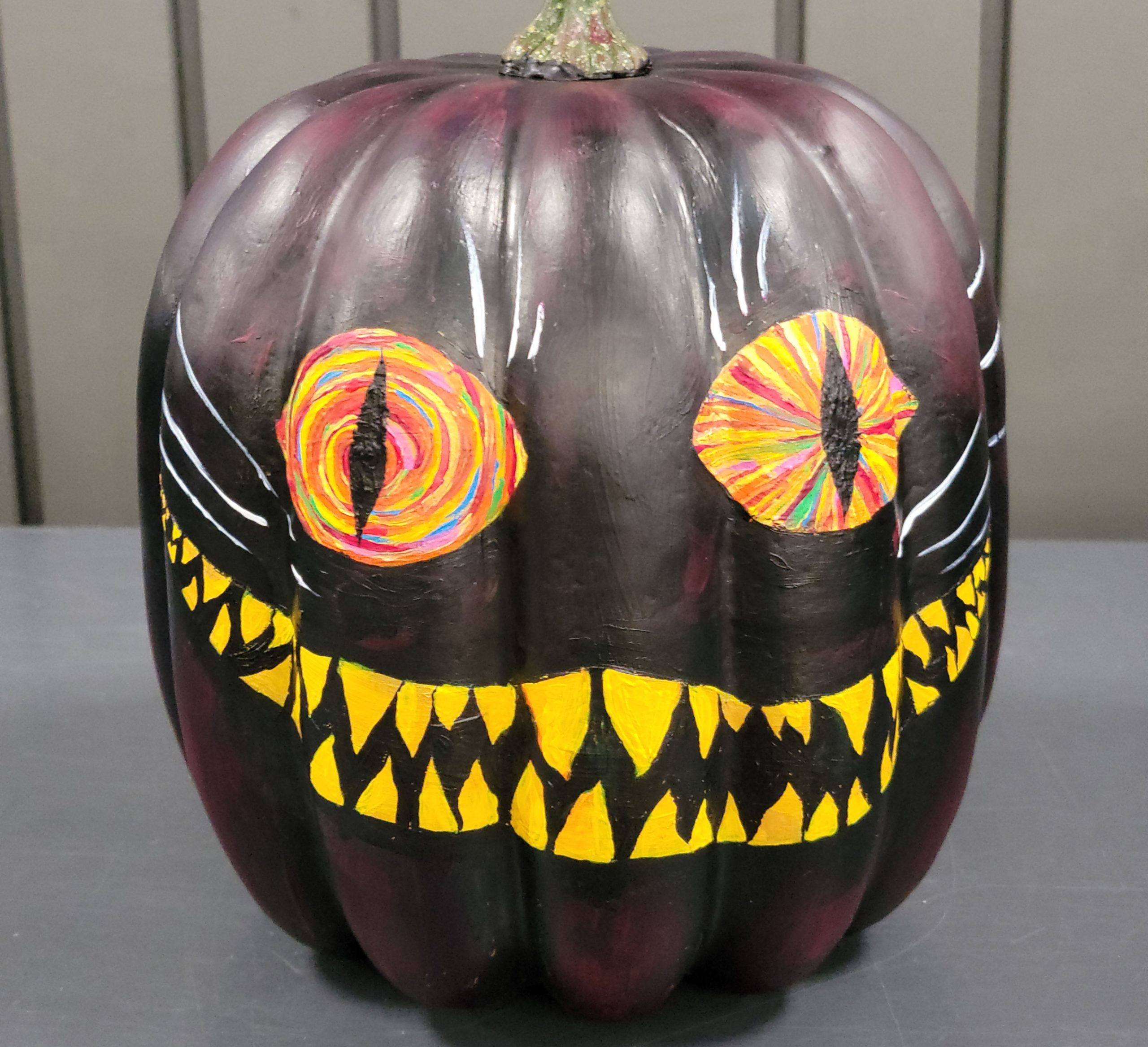 A pumpkin painted to look like the Cheshire Cat with a dark purple background and glowing orange eyes.