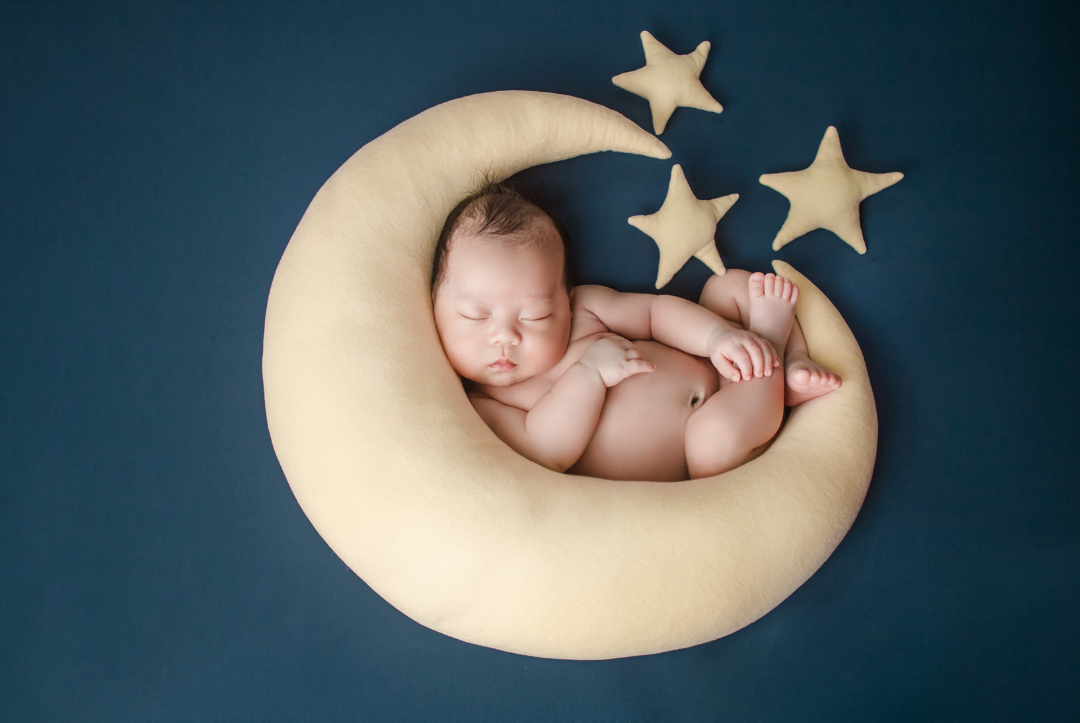 newborn baby sleeps on a crescent moon shaped pillow on a navy background