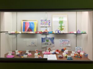 Display case of art projects including drawings, clay, and pipe-cleaner art
