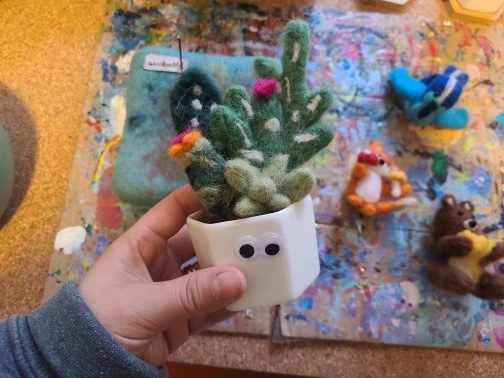 ID: A hand holds a felted succulent in a white container with googly eyes on it. In the background are more felting crafts.