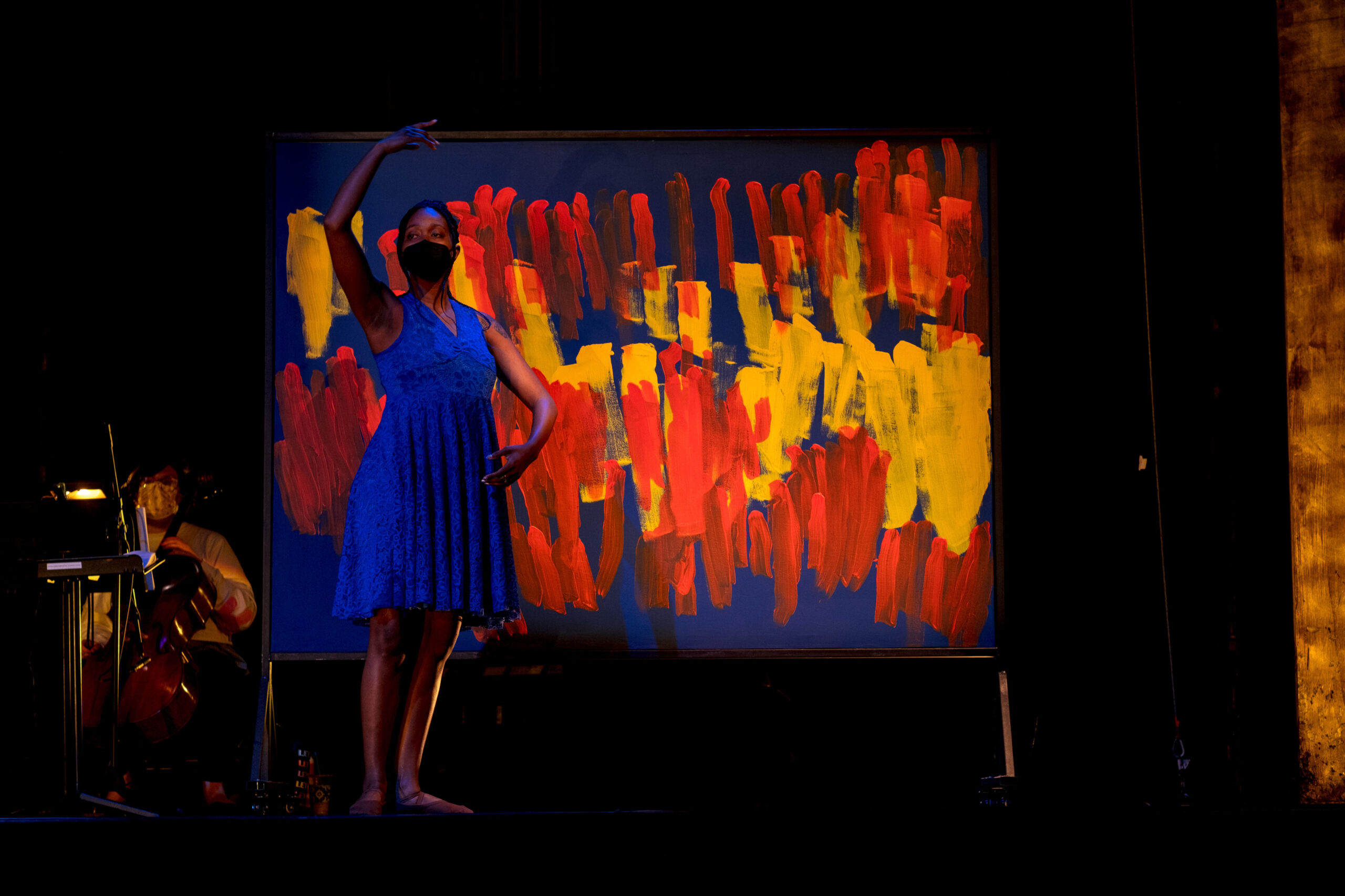 Image description: A Black woman in a facemask and blue dress stands in a dance pose with one arm above her head and the other in front of her. She is on a darkened stage, in front of a blue panel with red and yellow streaks on it.