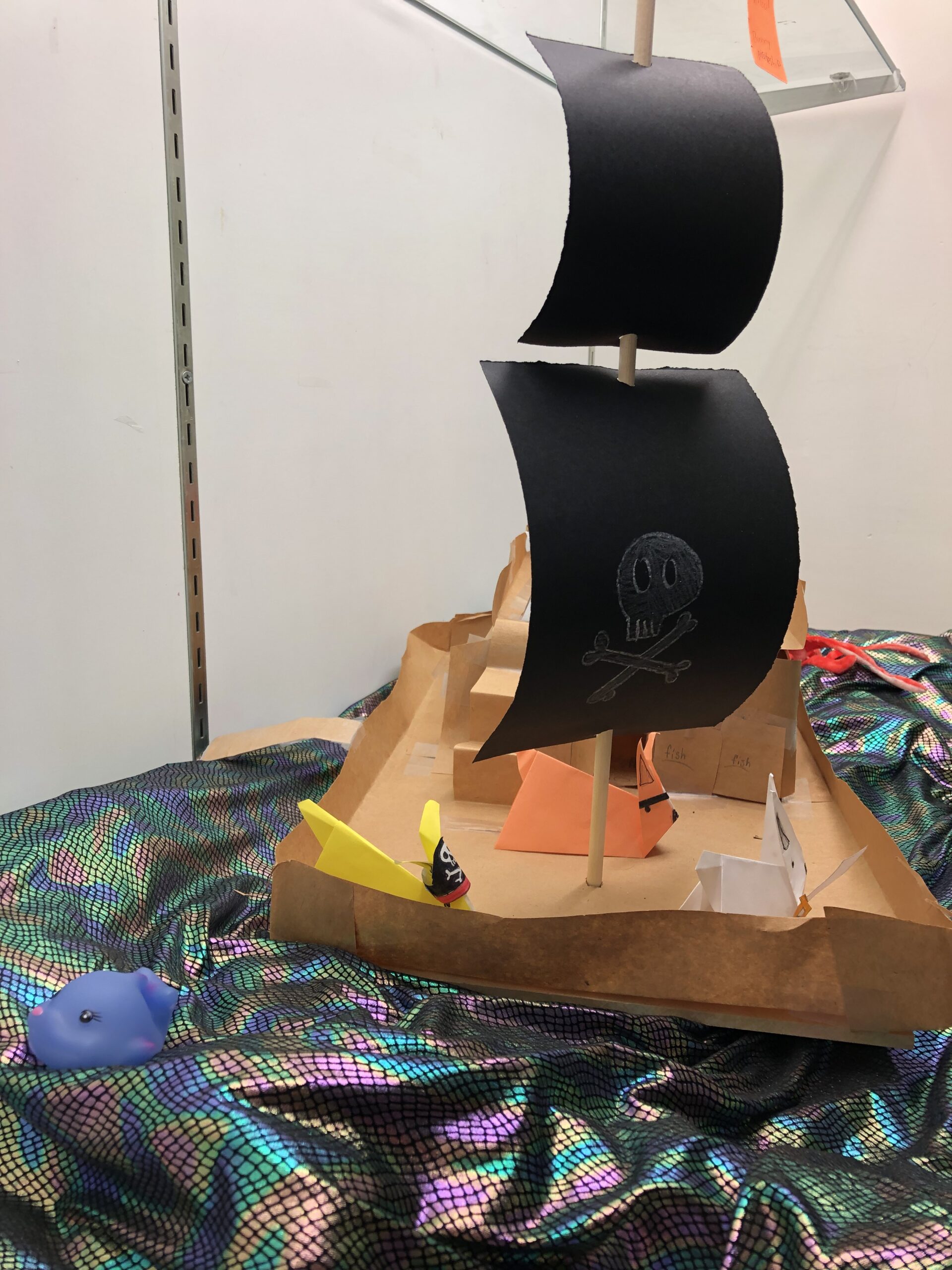 Pirate ship made of cardboard and paper, on top of a shiny, multicolored fabric mimicking waves. There are origami bunnies dressed like pirates aboard the ship.