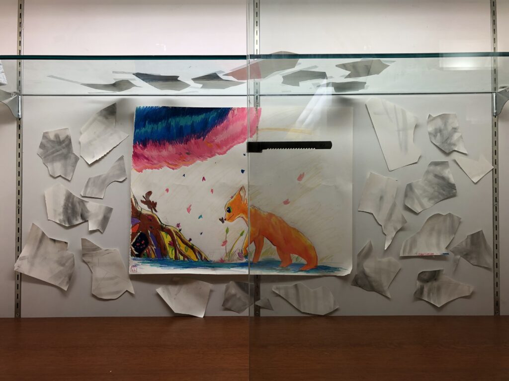 Display case with drawing of cat, tree, and cloud-formations, surrounded by broken gray pieces of paper.