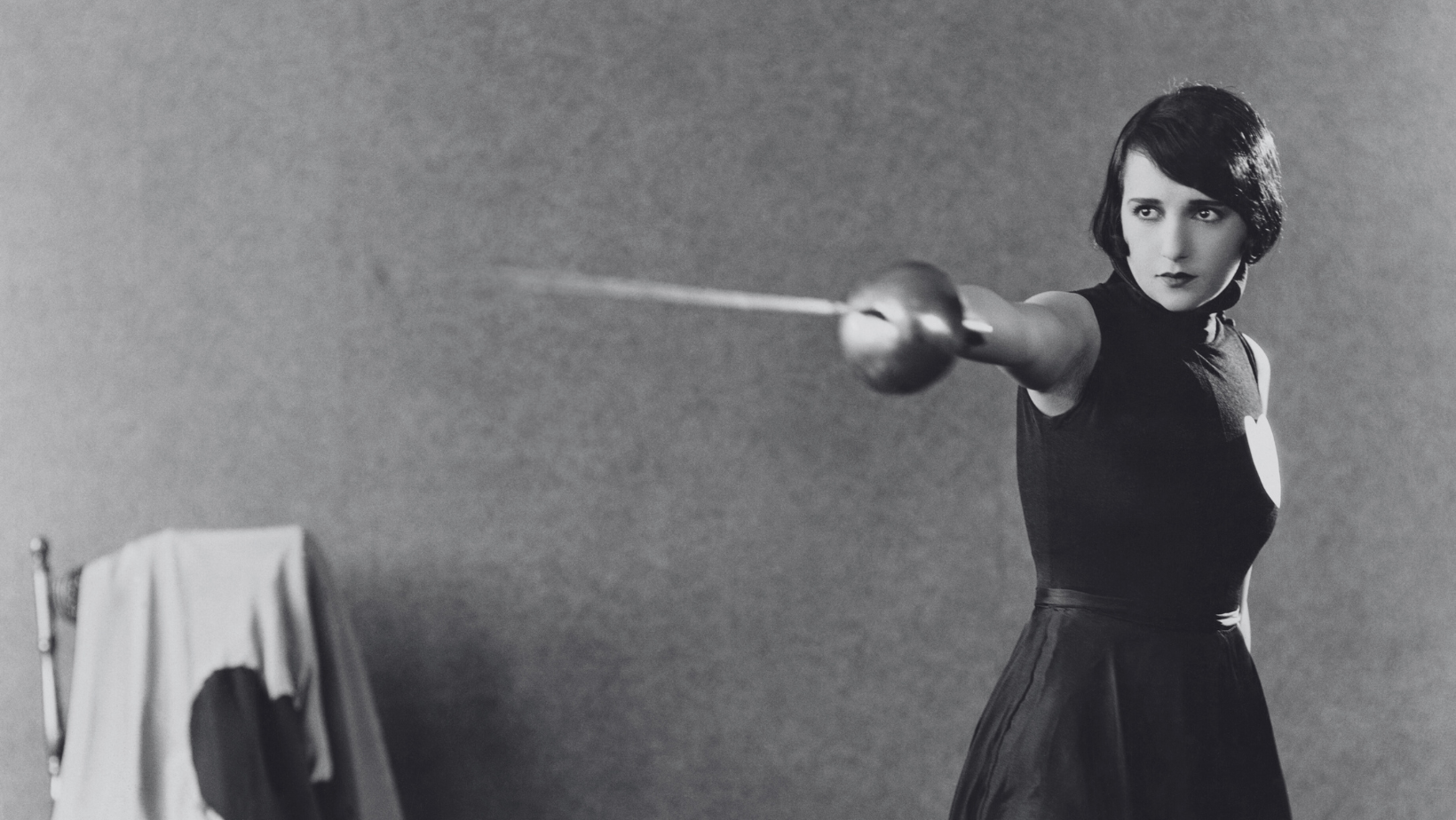 ID: A black and white picture of a short-haired woman pointing a sword.
