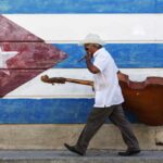 A musician carrying an upright bass in front of a mural of the Cuban flag