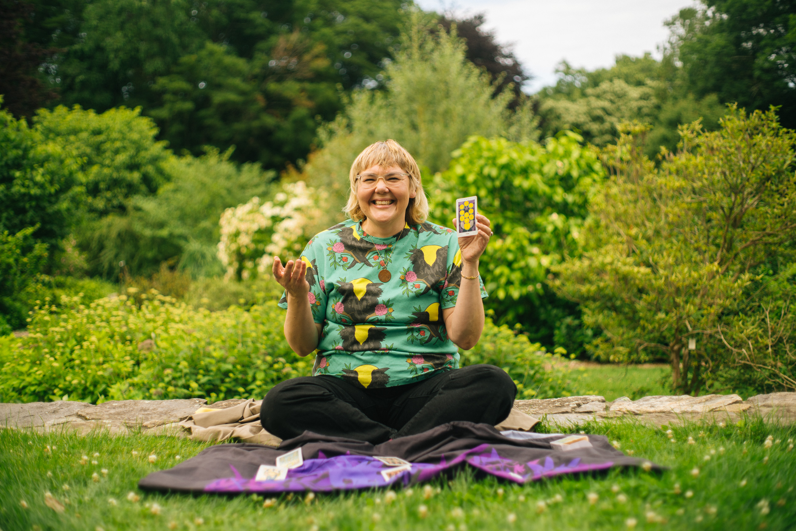 Person holding tarot cards and smiling, sitting on a blanket outside with greenery and bushes in the background