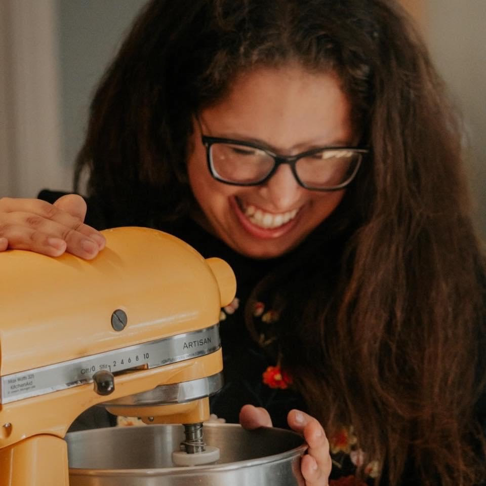Photo of a light-skinned woman with long brown hair and glasses, with a stand mixer.