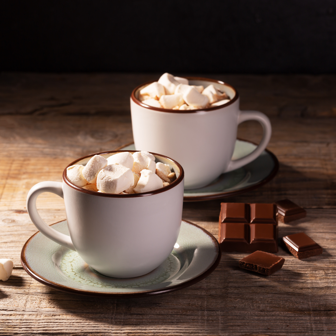 two mugs of hot chocolate with marshmallows, sitting on tea cup saucers, with a bar of chocolate on the side