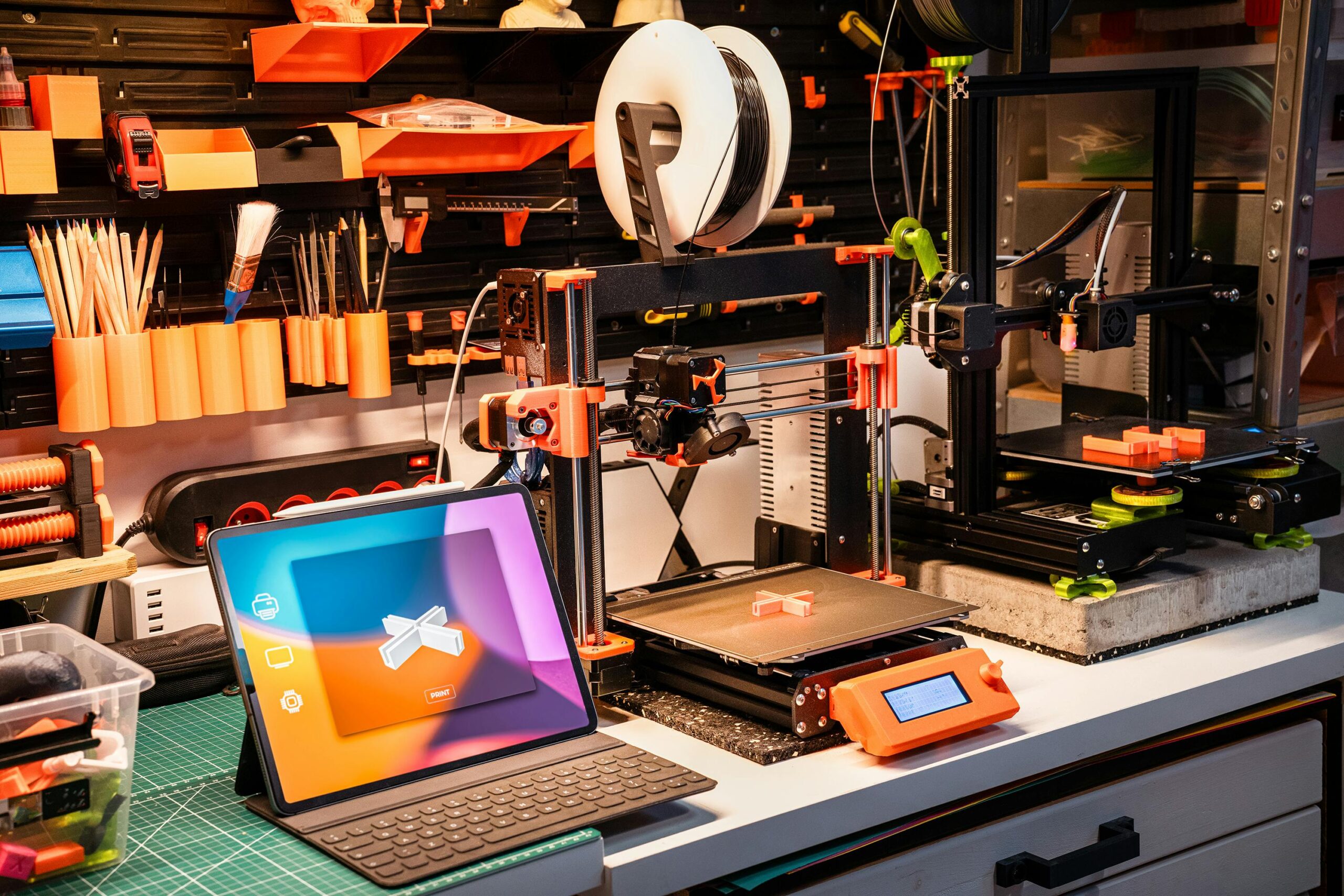 A workbench with a tablet and two 3D printers. The tablet displays a 3D plus sign, and one of the printers has printed the same design