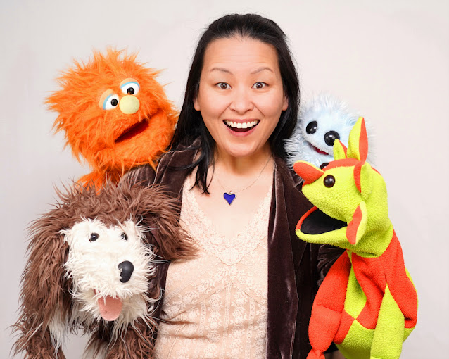 A dark-haired woman stands with an open-mouthed smile projecting surprise and delight. She is surrounded by four puppets: a green and orange puppet on her left hand, a small fuzzy blue creature on her left shoulder, an orange Muppet style puppet on her right shoulder, and a dog beside her right hand.