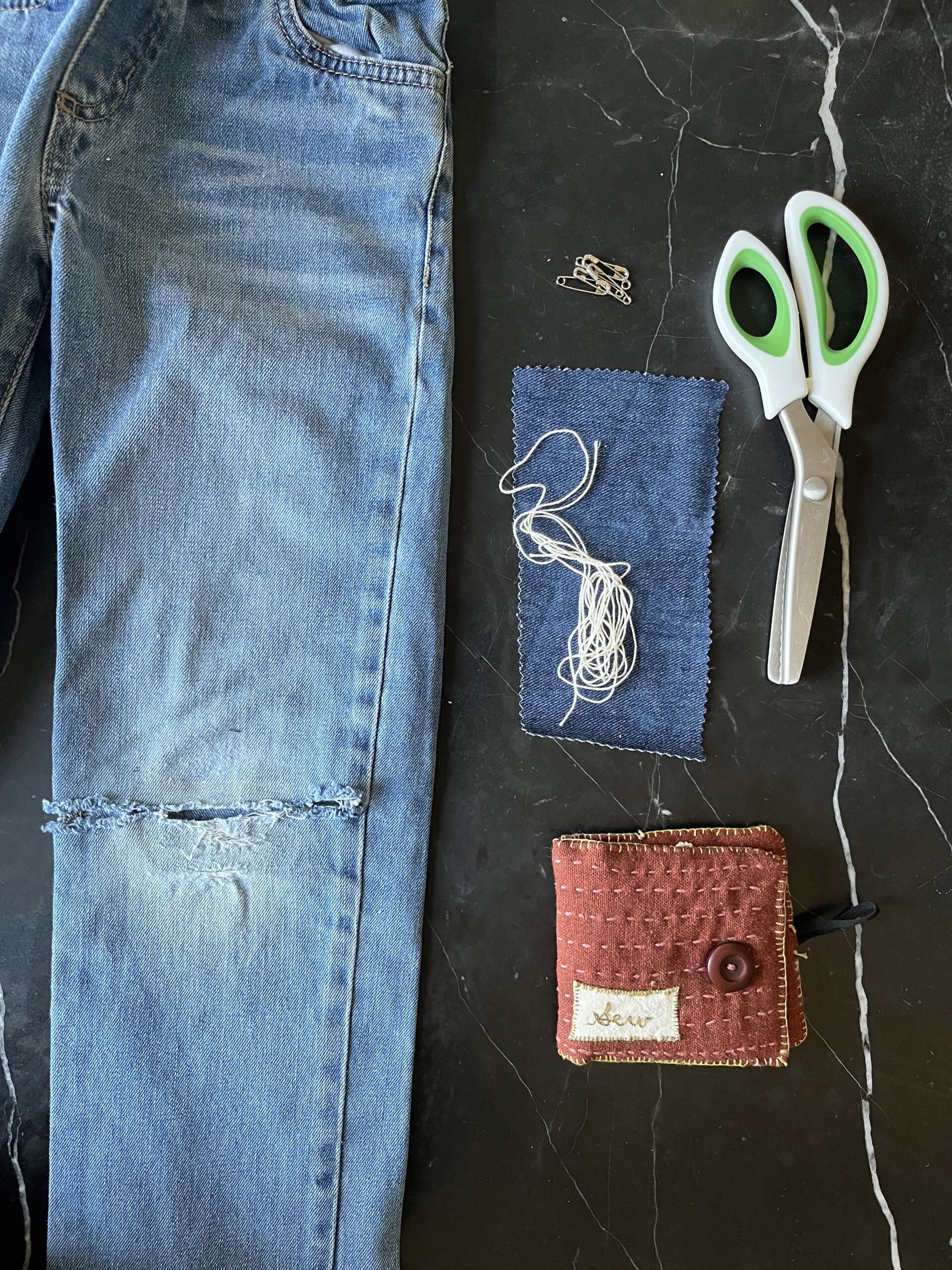 Image of a pair of denim pants with a damaged knee and the materials used to repair it: scissors, cloth, thread.